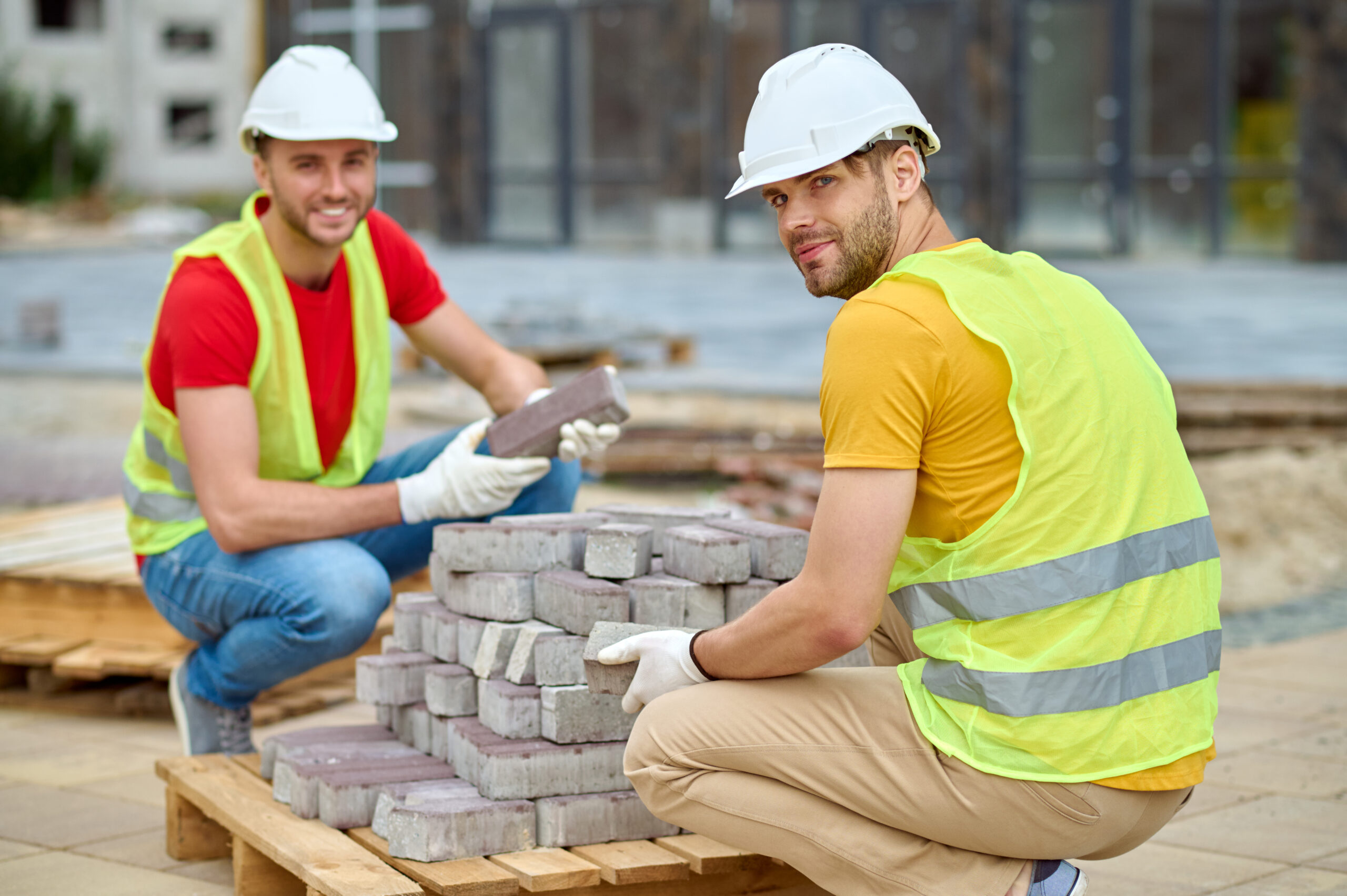 Manual labour. Two young adult smiling workers in protective helmet and gloves crouching stacking bricks looking at camera at construction site on warm day